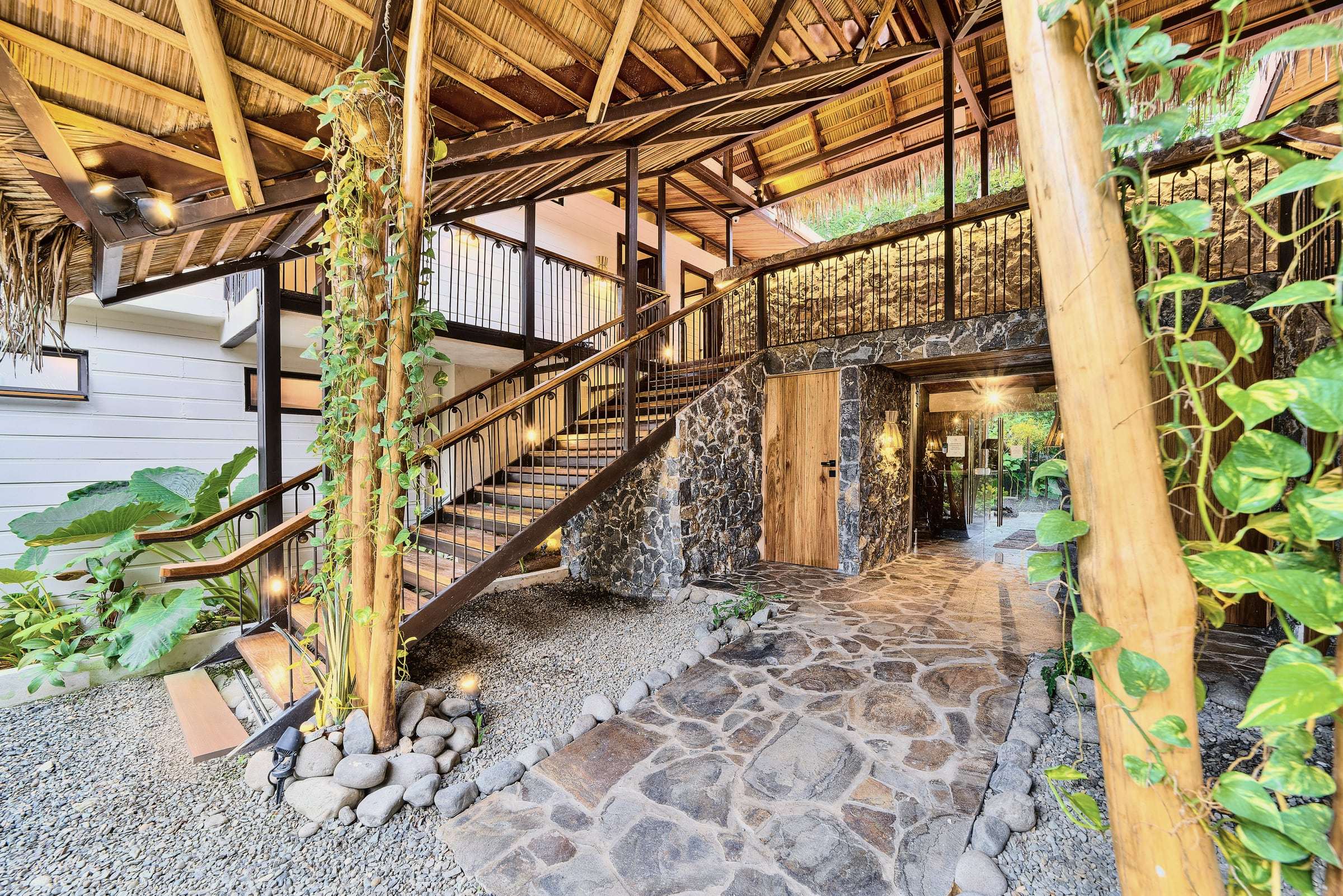 Atrium with vine-wrapped posts, an open wooden stair, stone walls and flagstones under the thatch