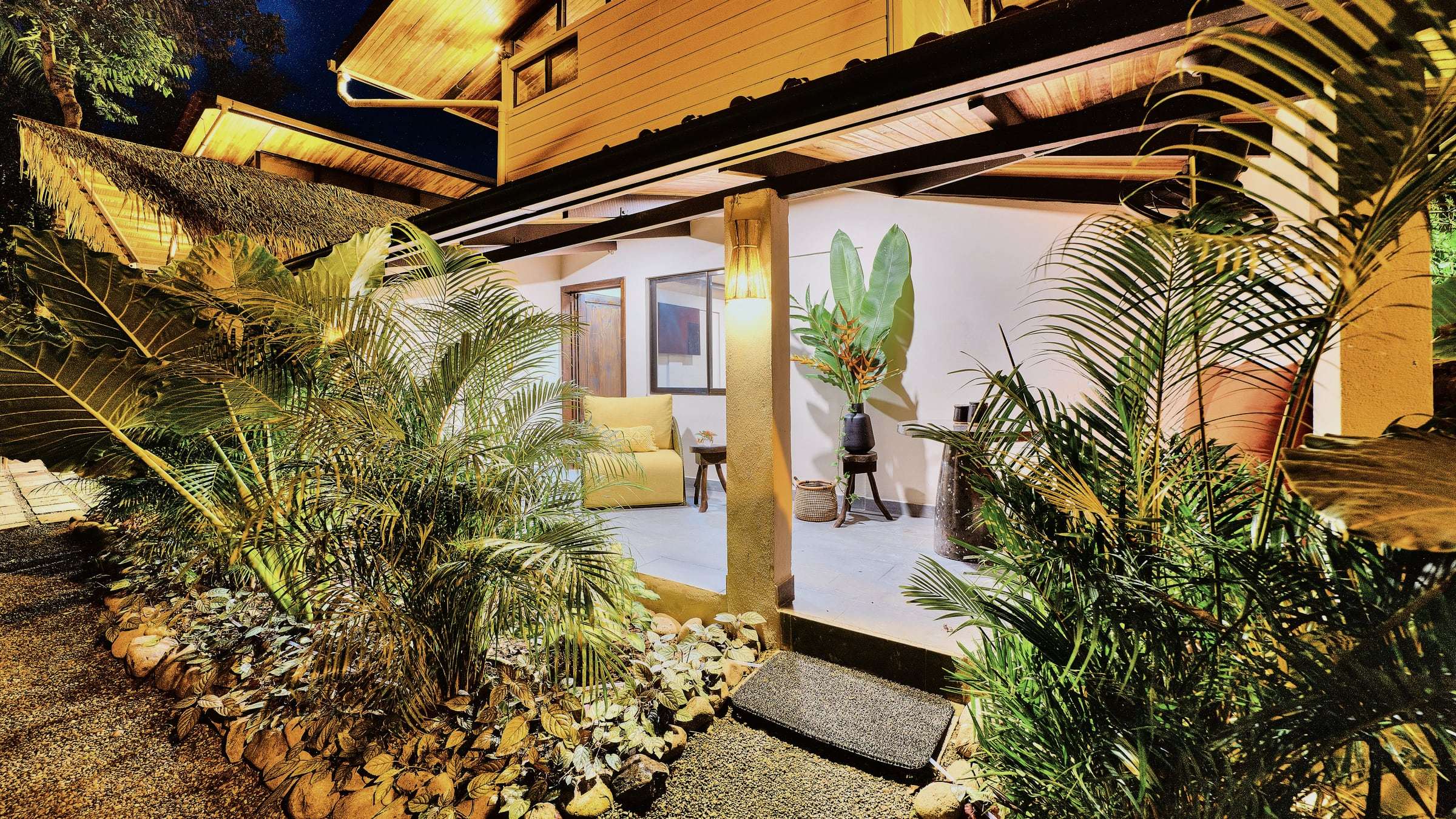 Ground-floor terrace at dusk: yellow armchair, tropical flowers and palms lit from below