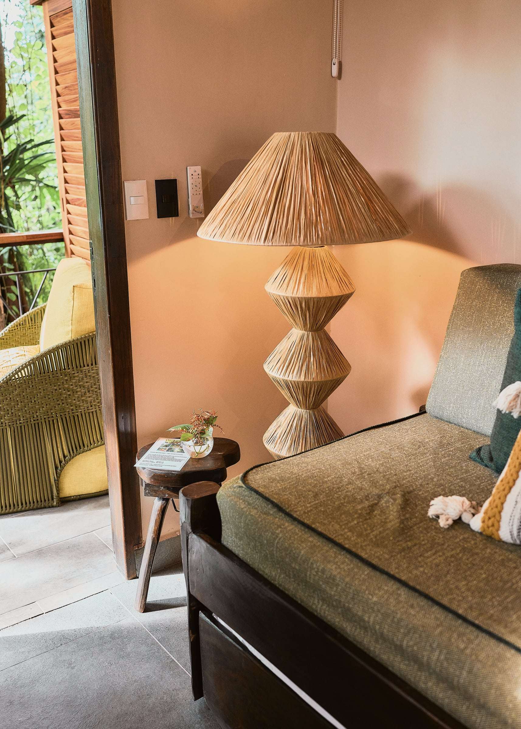 Raffia floor lamp beside a green daybed, with a yellow armchair on the terrace through the open door