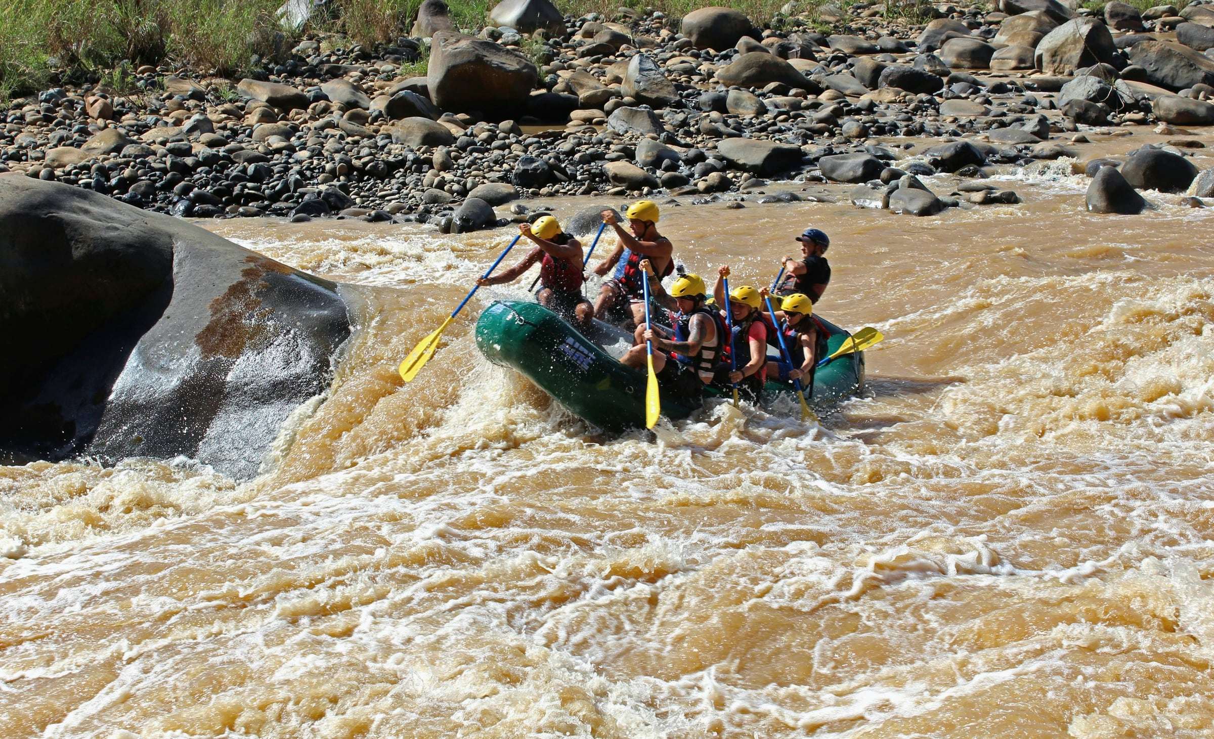 Rafters in helmets paddling a green raft through muddy white-water rapids on a Costa Rican river